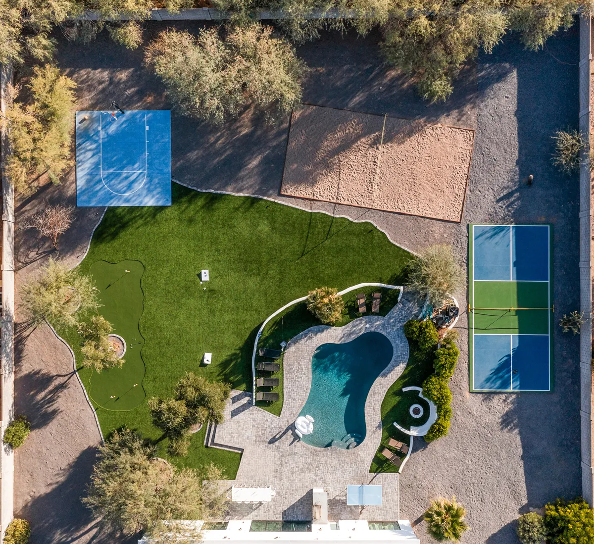 An expansive aerial view of the backyard showcasing a sparkling swimming pool, tennis court, basketball court, and sand volleyball area, all bordered by lush greenery. Multiple recreational zones are visible, seamlessly blending various amenities in one vibrant outdoor space.