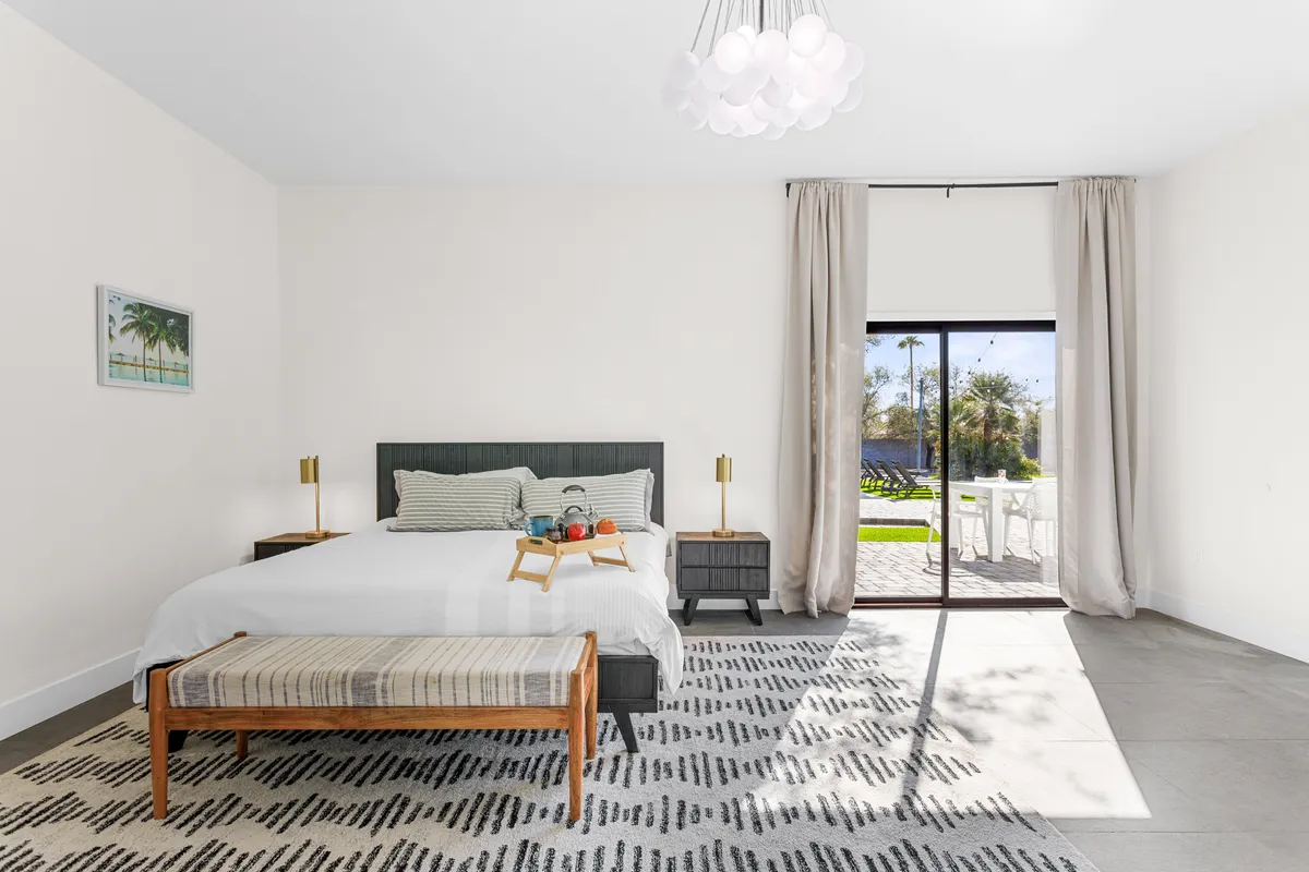 A tranquil bedroom featuring a plush bed, large window allowing for abundant natural light, a soft bench at the end of the bed, and a stylish light fixture overhead. The serene atmosphere is enhanced by soft tones and inviting decor that encourage relaxation and rest.