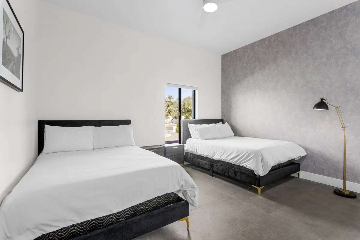 This image features a cozy bedroom with two beds, a large window letting in abundant natural light, and a floor lamp. The relaxed atmosphere is perfect for both families and groups, showing flexible sleeping arrangements ideal for both children and adults.