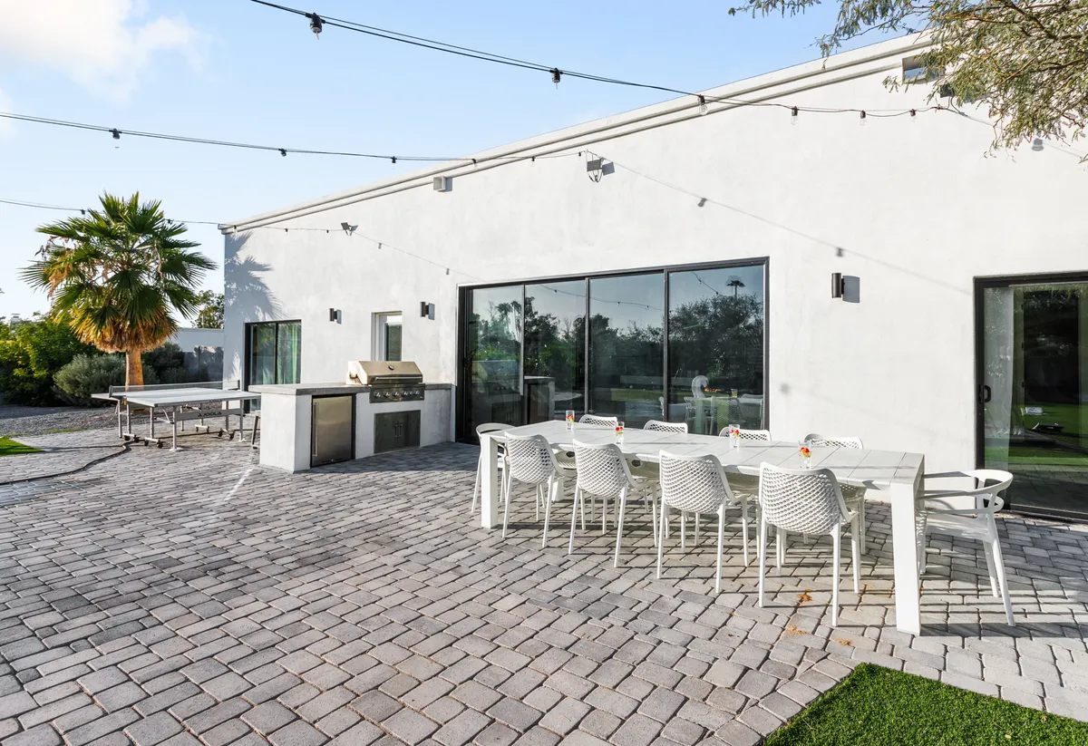 An inviting outdoor patio features a large dining table set for a meal, a built-in grill, palm trees, and spacious paved areas for socializing. String lights and the adjacent house’s large windows add a festive, relaxed ambiance perfect for evening gatherings and barbecues.