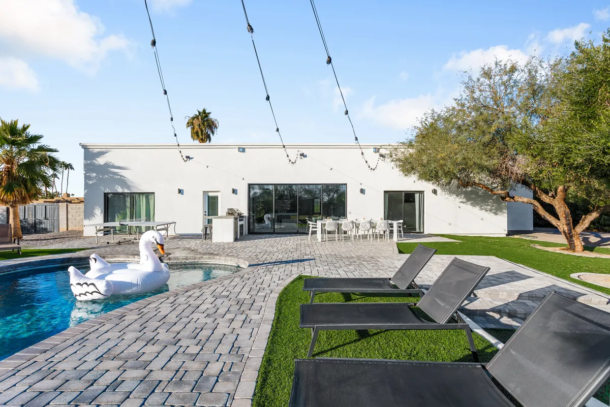 A cheerful outdoor scene featuring a sparkling swimming pool, comfortable lounge chairs, a decorative swan float, an outdoor dining area, and lush landscaping. The setting is perfect for sunbathing, swimming, or gathering with friends for meals and relaxation.