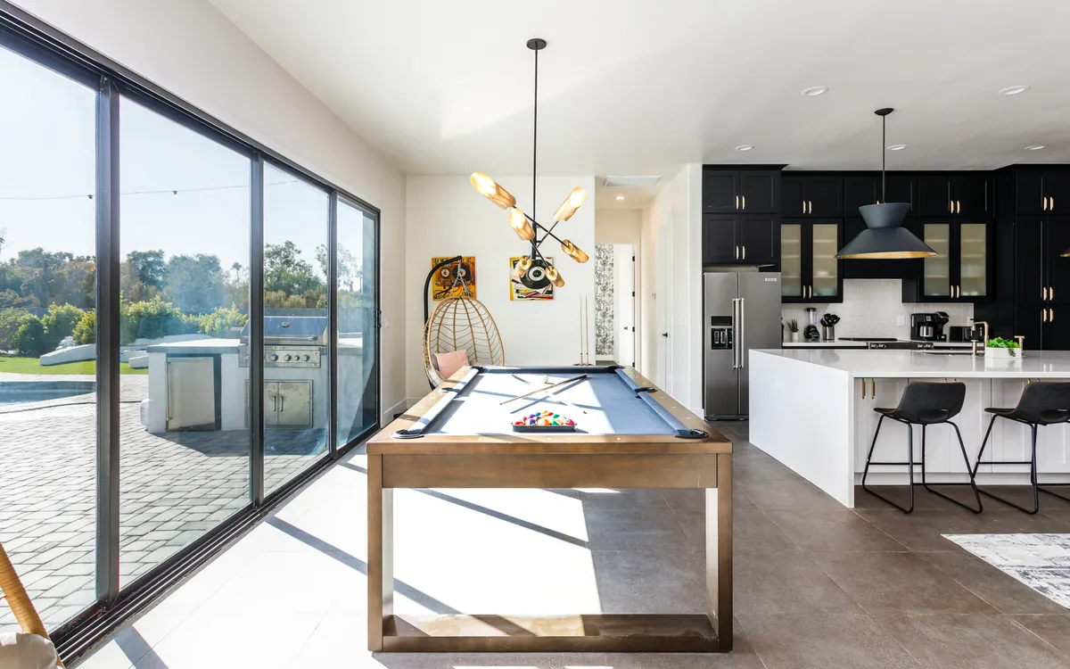 A lively recreation area highlighting a modern pool table set in a bright room with large windows, stylish lighting, and contemporary artwork. The adjacent spaces suggest an open layout that encourages socializing and interactive fun.