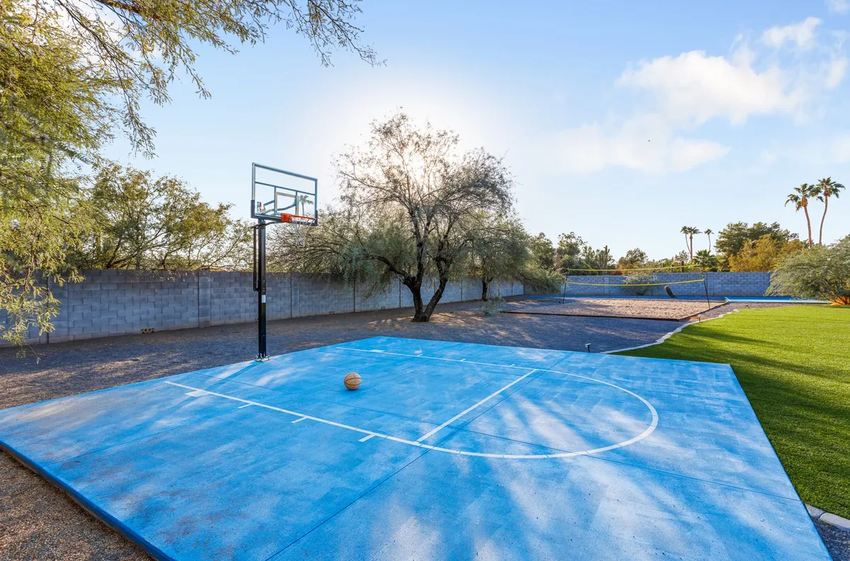 This exterior shot captures a vibrant blue basketball half-court surrounded by lush green grass and trees, with a hoop ready for action. The area is bathed in sunlight, creating an energetic and playful outdoor atmosphere ideal for group sports and gatherings.