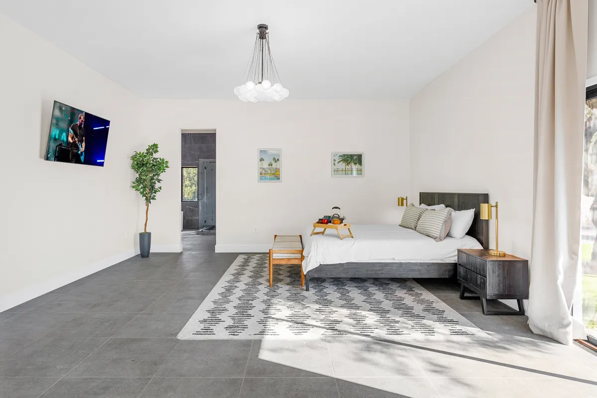 A serene and comfortable bedroom with a cozy bed, elegant rug, side table, a lush plant for a touch of nature, and a wall-mounted television. The room exudes a warm, inviting feel, perfect for relaxing, reading, or watching TV.